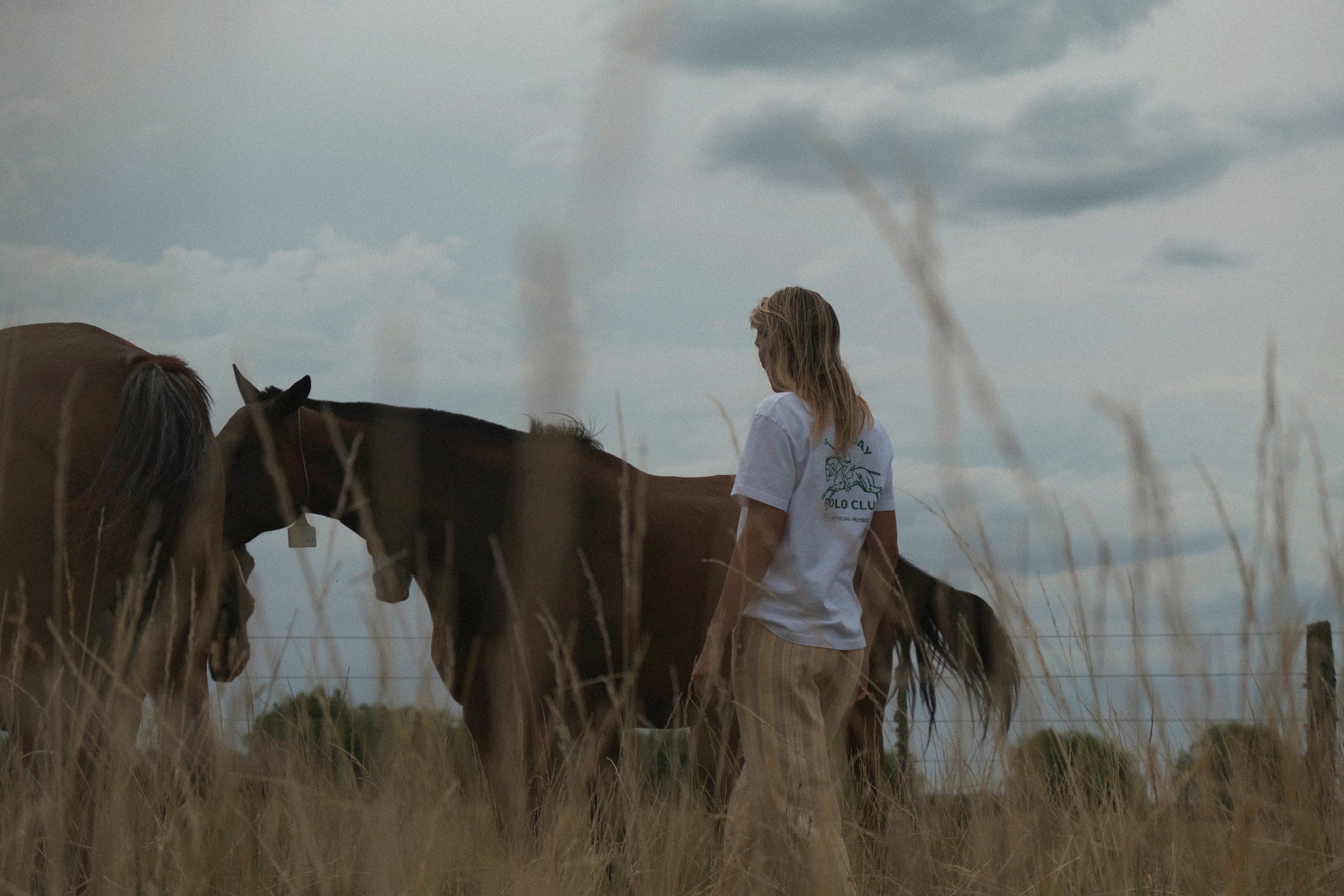 Person standing wearing 'Sunday Polo Club' tee with two horses in a field with a cloudy sky.