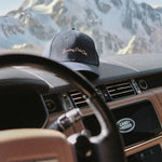 Black cap with 'Sunday Polo Club' text on a car dashboard with a mountain view in the background