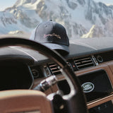 Black cap with 'Sunday Polo Club' text on a car dashboard with a mountain view in the background